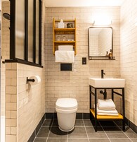 Un baño moderno con inodoro, lavamanos y espejo. Hay repisas con artículos de aseo y un perchero.