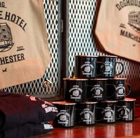 Una pila de tazas negras con el logotipo del Doghouse Hotel Manchester, sobre una superficie de madera con fondo de malla y una bolsa de toallas del Doghouse Hotel Manchester.