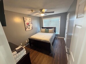 A bedroom with a bed, a ceiling fan, a window with blinds, a painting on the wall, and a door. - Beautiful Private 3 Bedroom Home (Albany)