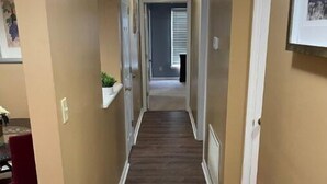 A hallway with wooden flooring, beige walls, and white trim. There are doors on both sides, and a plant is visible on the left wall. - Beautiful Private 3 Bedroom Home (Albany)