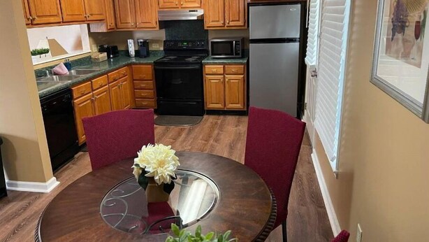 A compact kitchen with wooden cabinets, a stainless steel refrigerator, a black stove, and a dining table with chairs. - Beautiful Private 3 Bedroom Home (Albany)