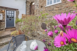 A stone building with a door and window, a chair, a table with a teapot and cups, and pink flowers.