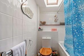 A bathroom with white tiles, a wooden toilet seat, a towel rack, a shelf with toiletries, a shower curtain, and a skylight.