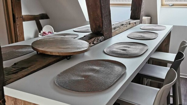 Una larga mesa de comedor con individuales tejidos, vigas de madera y sillas blancas en un interior rústico.