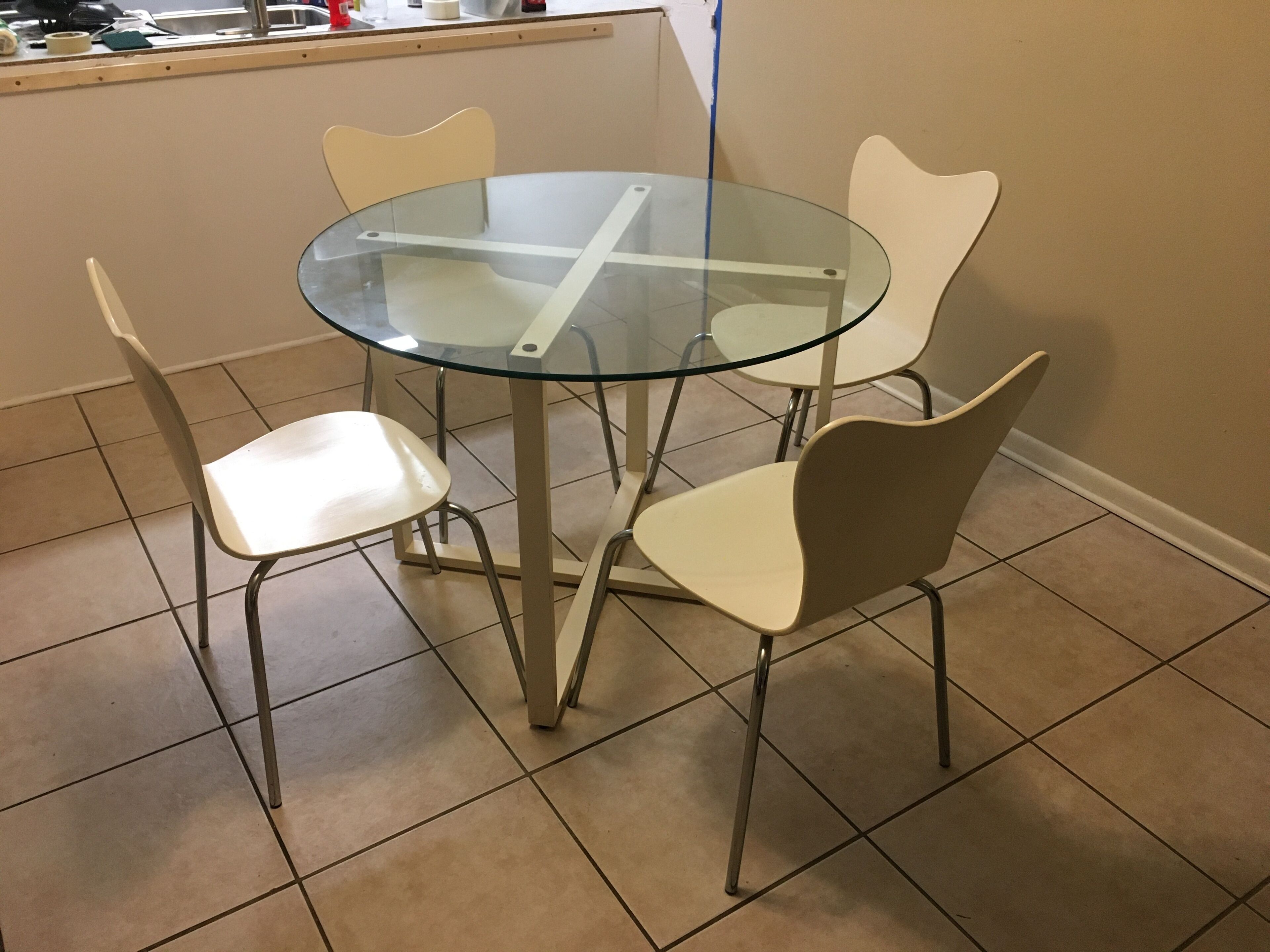 Ein runder Glastisch mit vier Stühlen, jeweils mit geschwungener Rückenlehne. Der Tisch steht auf Metallbeinen. Der Boden ist mit quadratischen Fliesen belegt.