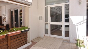 Entrada de hotel com um banco de madeira, plantas em vasos e uma porta de vidro que dá para o interior.