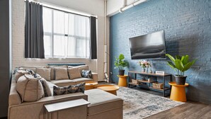 A modern living room with a sofa, ottoman, and a flat-screen TV mounted on a brick wall. - *New* Huge!  Spacious 3 Bedroom in Oakland that sleeps 12! over 3000+ sq ft (Pittsburgh)