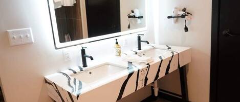 Un baño con tocador blanco, grifos negros y un espejo grande. Hay un perchero en la pared y una puerta a la derecha.