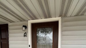 Una puerta de madera con un panel de vidrio, un buzón marrón oscuro y un felpudo negro en un porche con revestimiento blanco.