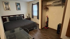 Un dormitorio con cama, escritorio, ventilador de techo, ventana con cortinas y armario con ropa colgada.
