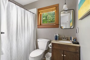 Een badkamer met toilet, een wastafel met toiletartikelen, een douchegordijn, een raam met uitzicht en een spiegel.