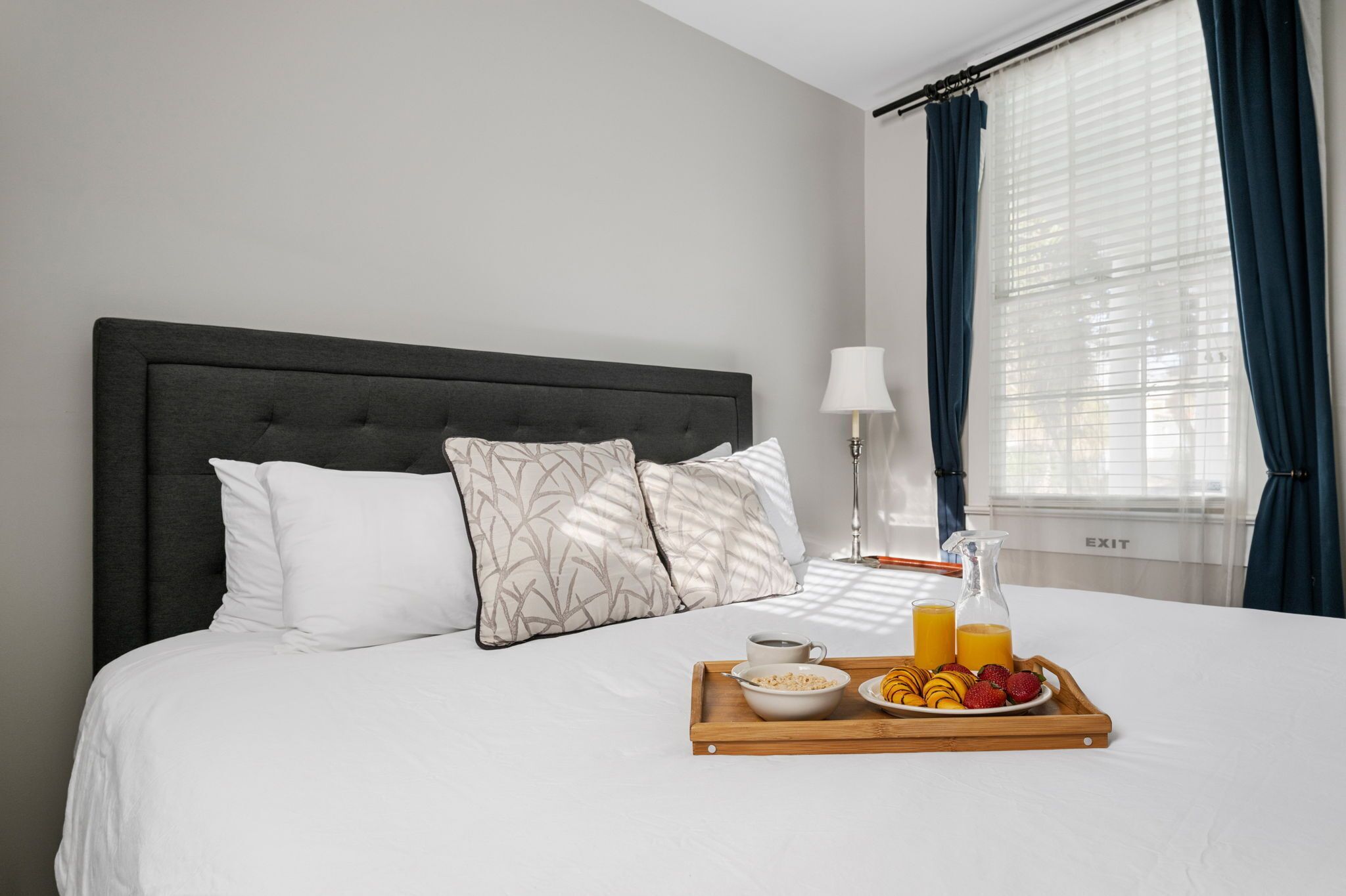 A neatly made bed with a tray of breakfast including cereal, fruit, and juice, a bedside lamp, and a window with curtains.