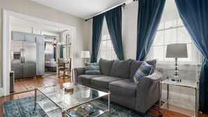 A modern living room with a grey sofa, a glass coffee table, and a kitchen area in the background. - Great Neighborhood Restaurants | Lewis Court - A (Charleston)