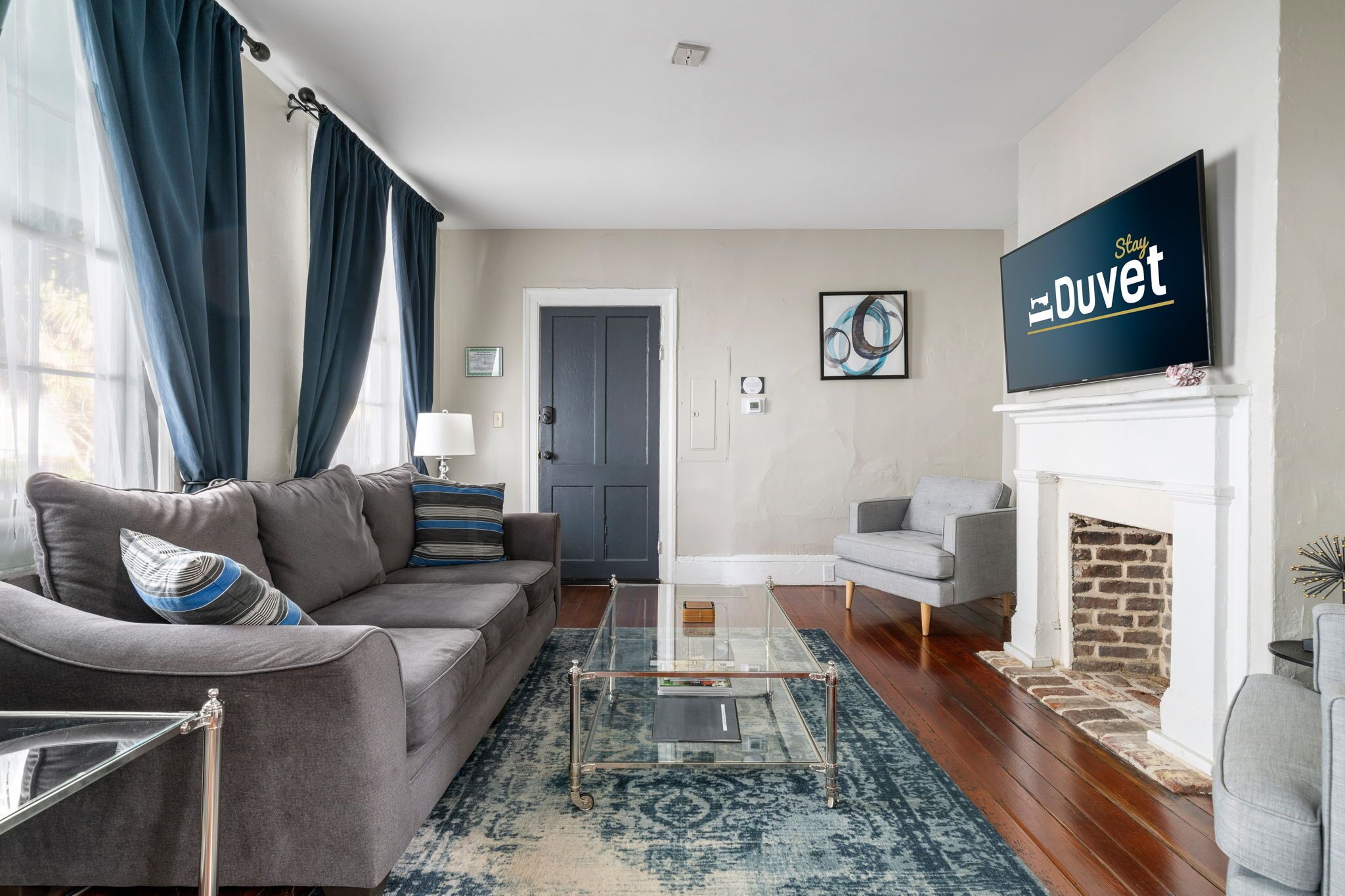 A living room with a gray sofa, a glass coffee table, a fireplace, and a TV on the wall.