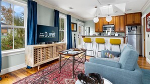 Un salon moderne avec un canapé, une table basse et un téléviseur posé sur un meuble en bois. Il y a également un coin cuisine équipé d’un four à micro-ondes, d’un réfrigérateur et de tabourets de bar.