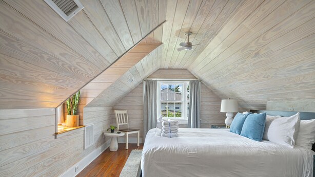 A bedroom with a sloped ceiling, a bed with white bedding, a small table with a lamp, and a window with curtains.