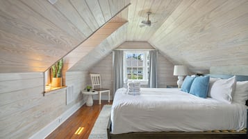 Ein Schlafzimmer mit schrägen Wänden, einem Bett mit weißer Bettwäsche, einem kleinen Tisch mit Lampe und einem Fenster mit Vorhängen.