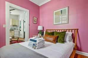 A hotel room with a bed, pillows, a bedside table with a lamp, a framed picture on the wall, and a view of the kitchen through an open door.
