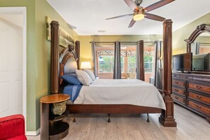 Uma cama de dossel com ventilador de teto, uma televisão em uma cômoda e vista para o exterior por uma porta de vidro.