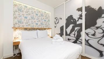 A neatly made bed with white linens and a folded towel, a wooden nightstand with a lamp, and a floral-patterned headboard.