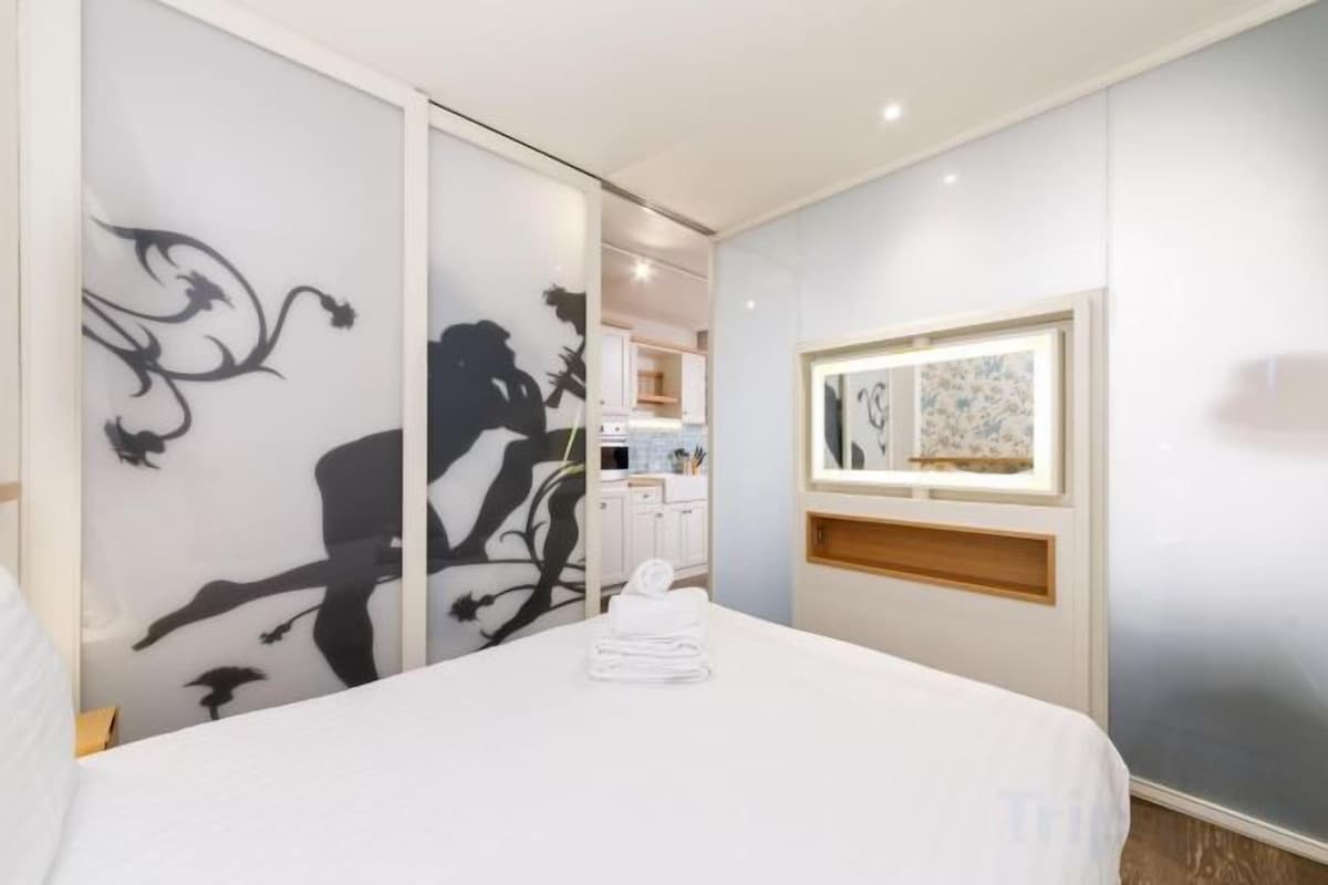 A hotel room with a bed, a sliding door with artwork, a window with a view, and a small kitchen area.