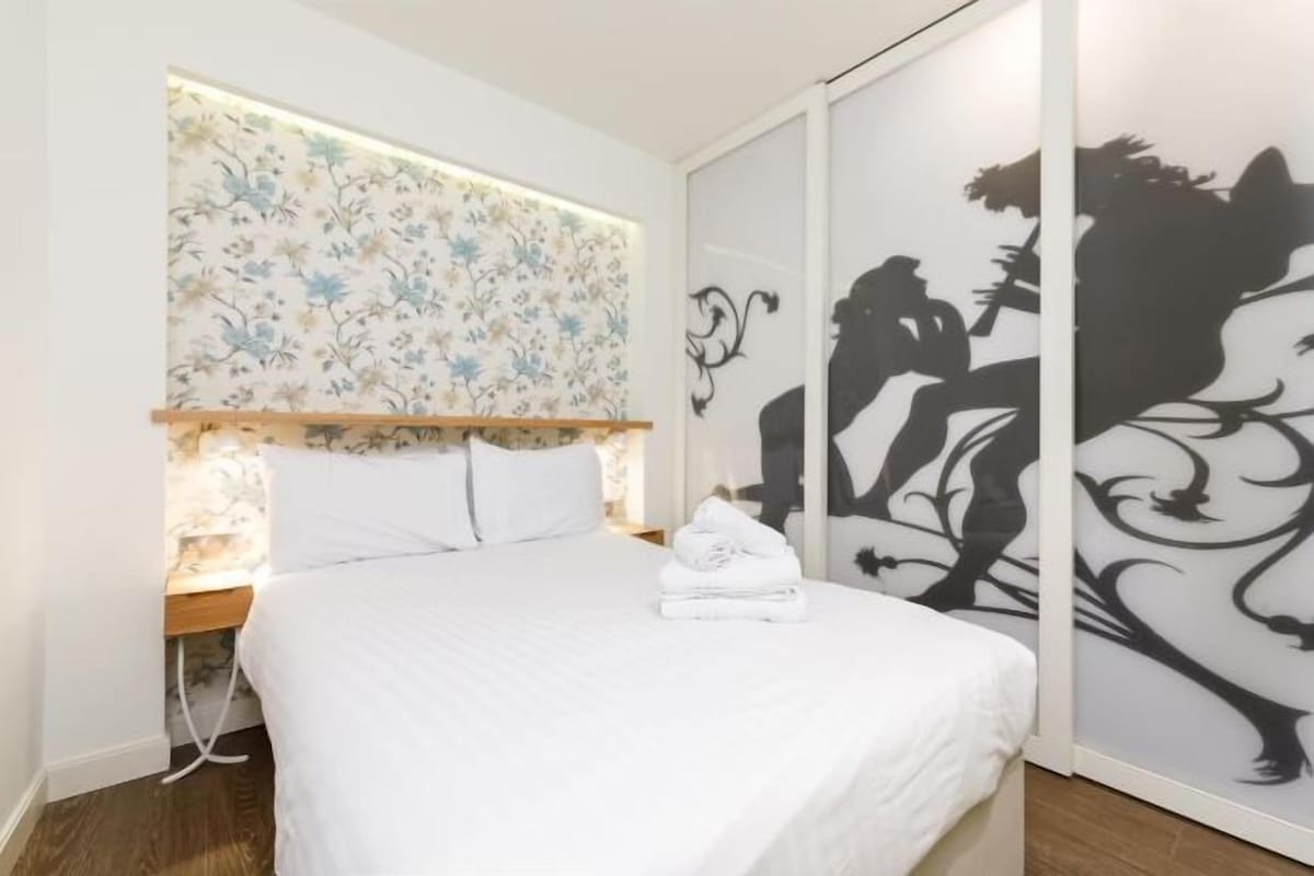 Une chambre à coucher comprenant un lit, une table de chevet, un mur recouvert d’un papier peint à motifs floraux et une silhouette décorative sur le mur.