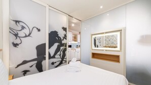 Ein Hotelzimmer mit Bett, einer Wand mit dekorativer Silhouette, einem kleinen Fenster mit Spiegel und Blick auf den Küchenbereich.