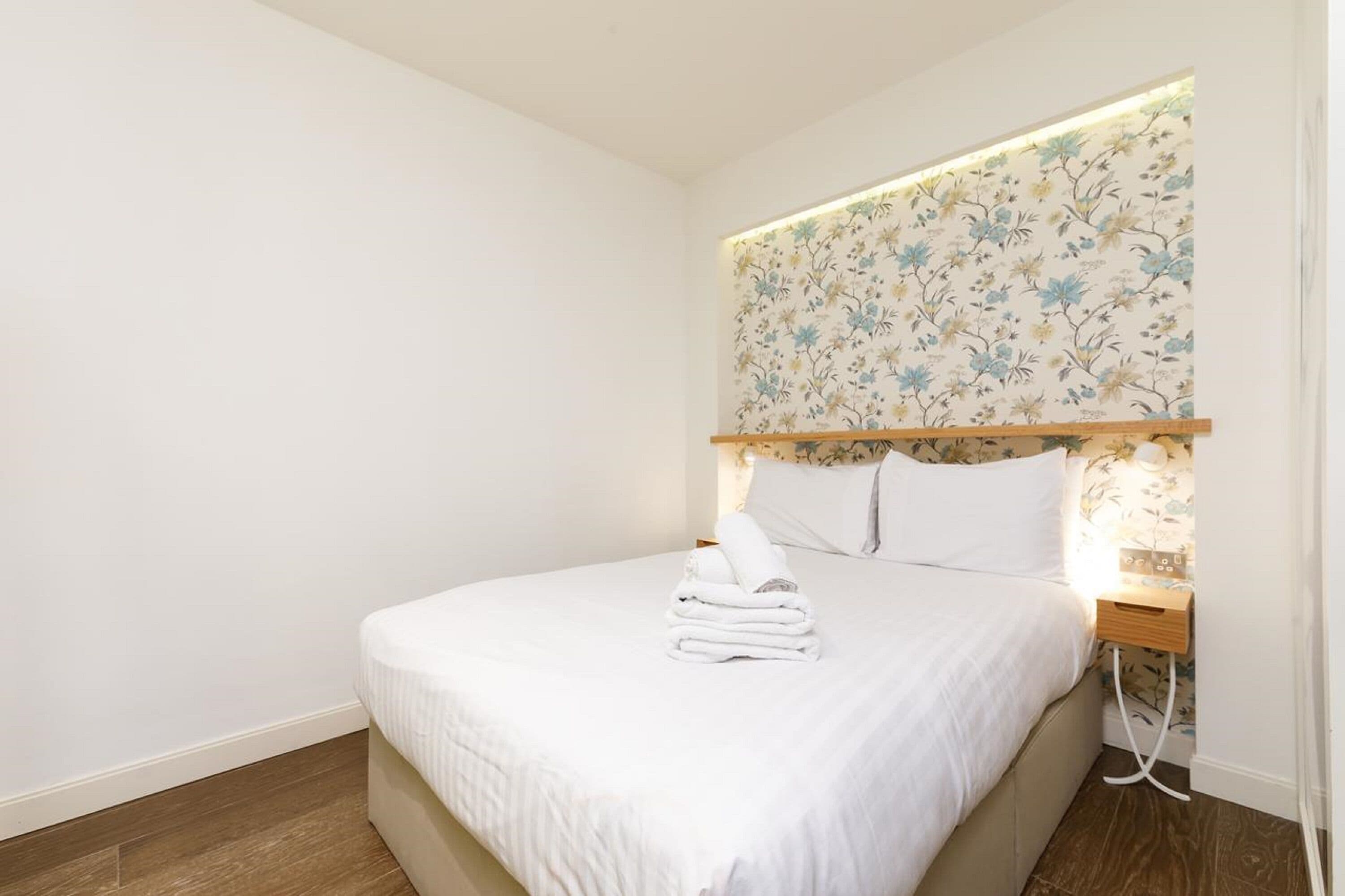 A neatly made bed with white linens and a stack of folded towels, a wooden headboard with floral wallpaper, and a bedside table with a lamp.