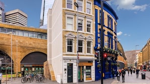 Straßenansicht mit einer Mischung aus moderner und traditioneller Architektur, darunter ein Gebäude mit einer Schaufensterfront und ein blaues Eckgebäude.