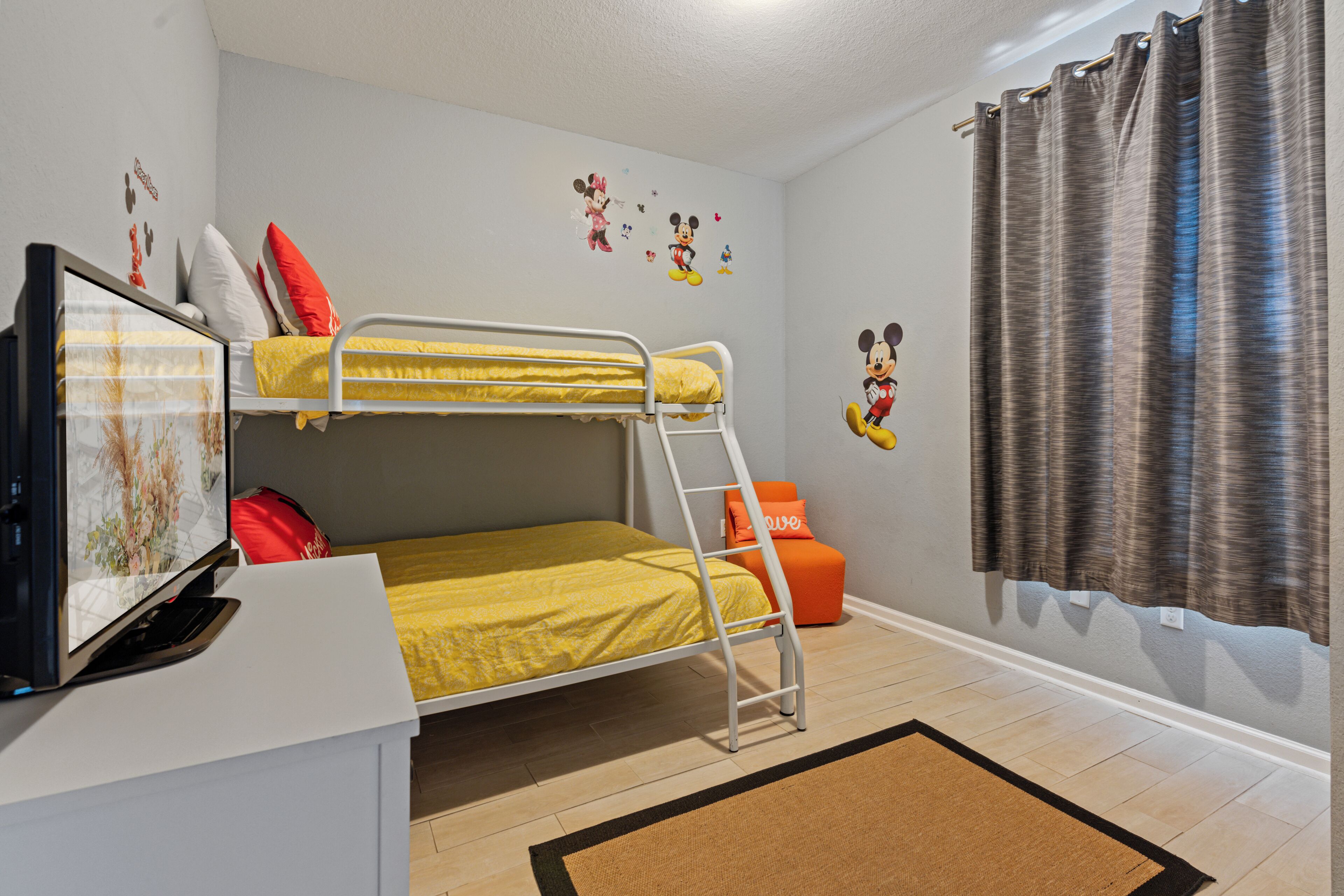 A bunk bed room with a television, Mickey Mouse wall decal, and a window with curtains.