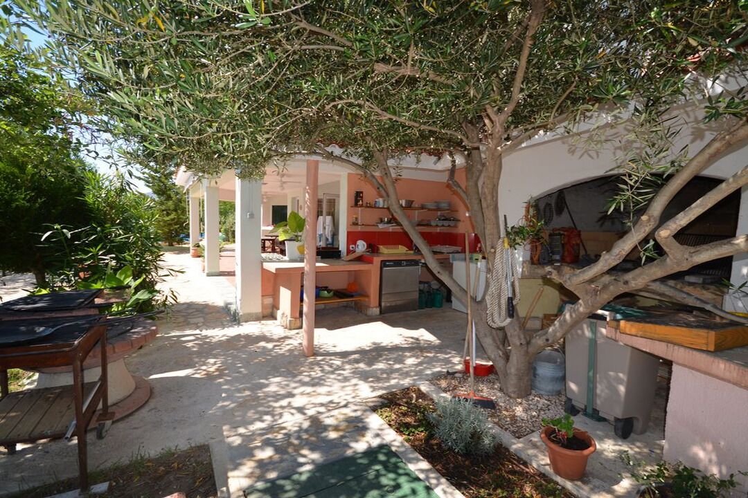 A covered outdoor area with a dining table and chairs, a small garden with potted plants, and a building with a kitchen and dining area visible through an archway.