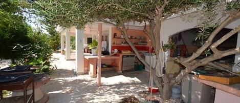 A covered outdoor area with a dining table and chairs, a small garden with potted plants, and a building with a kitchen and dining area visible through an archway.