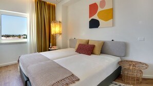 A double bed with a beige and white bedspread, two pillows, a woven side table, a wall art piece, and a window with curtains. - Boavista Apartment - Vintage & romantic! (Albufeira)