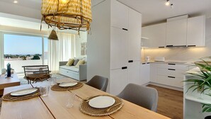 A modern kitchen with white cabinets, a dining area with a wooden table set for four, and a view of the outdoors through large windows. - Boavista Apartment - Vintage & romantic! (Albufeira)
