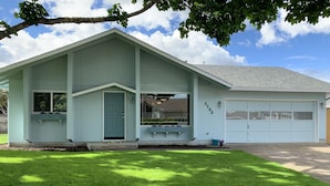 Une maison d’un seul étage, de couleur vert clair, avec un garage, un porche à l’avant doté d’un banc et une pelouse bien entretenue.