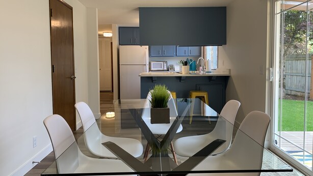 Une cuisine moderne avec des meubles de couleur bleu foncé, un réfrigérateur blanc et un four à micro-ondes. On y trouve une table à manger en verre, quatre chaises blanches et une petite plante en pot.