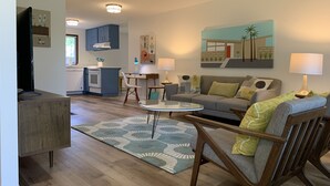 Un salon moderne comprenant un canapé, des fauteuils, une table basse et un coin repas avec une table et des chaises. Au fond, on aperçoit une cuisine avec des placards et un plan de travail.