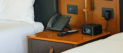 A hotel room with a bed, a bedside table with a lamp, a phone, and a digital clock.