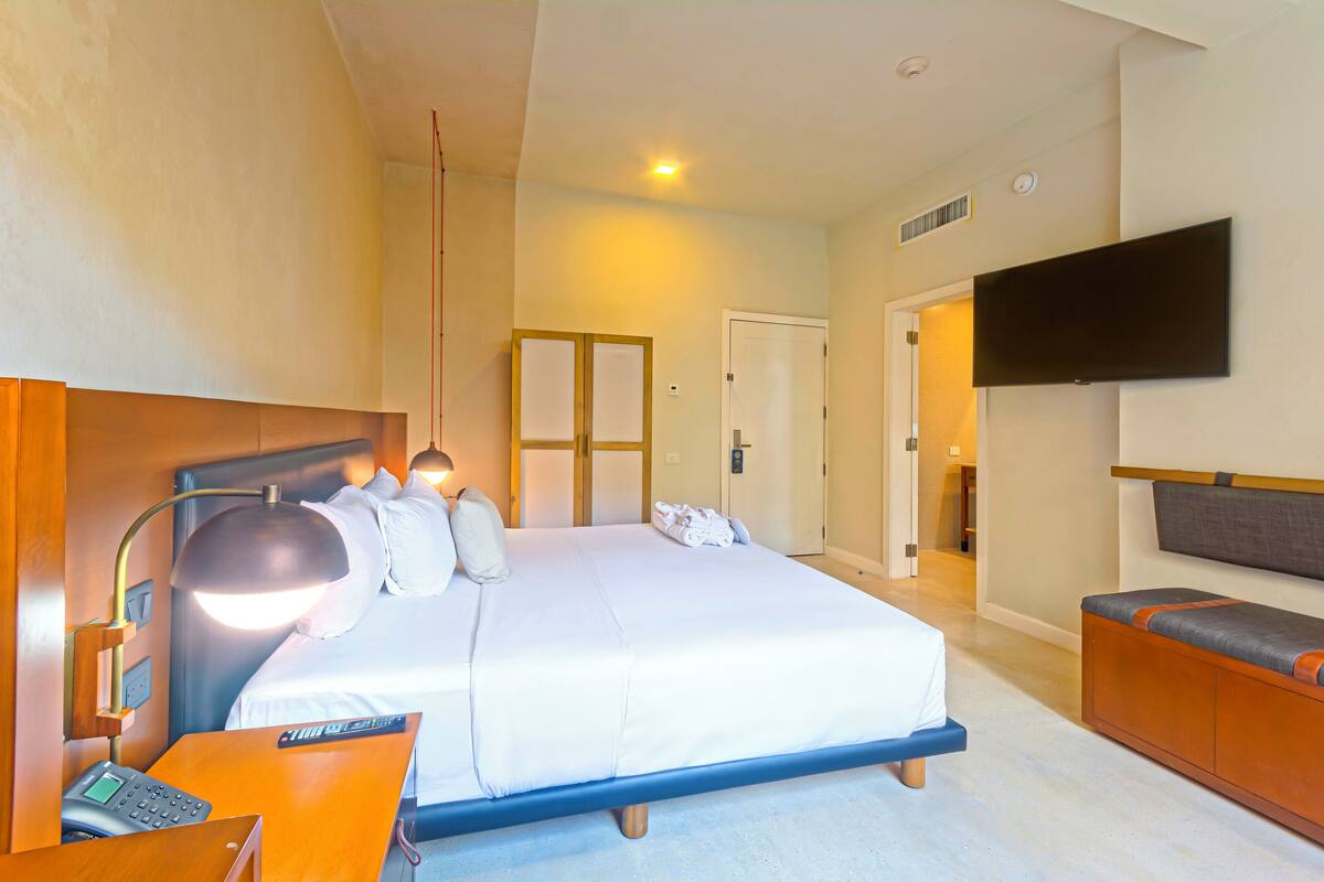 Une chambre d’hôtel avec un grand lit, une lampe de chevet, un téléviseur à écran plat fixé au mur, un banc en bois et une porte donnant sur une autre pièce.