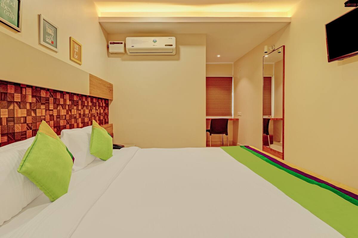 A hotel room with a wooden headboard, a bed with white linens and green pillows, a flat-screen TV mounted on the wall, and a large mirror.