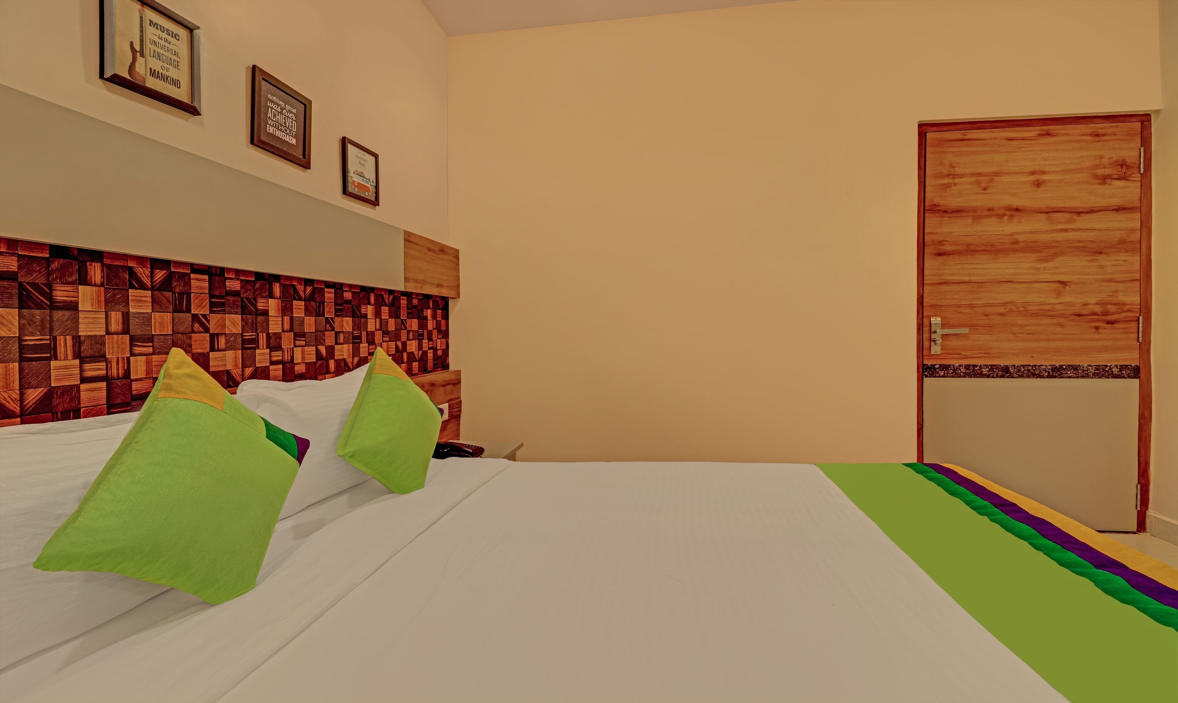 A hotel room with a wooden headboard, a bed with white bedding and green pillows, a wooden door, and two framed motivational quotes on the wall.