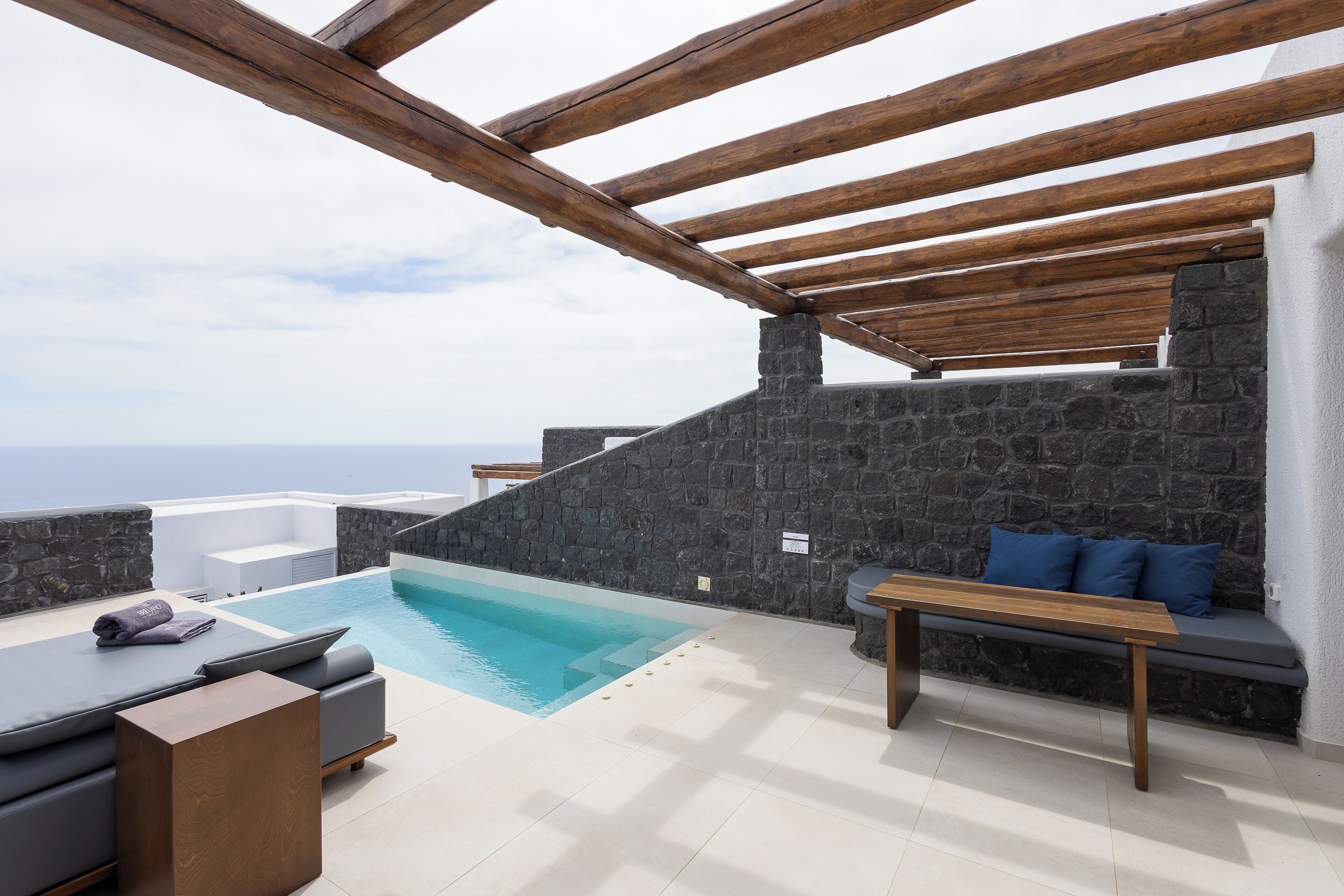 Área de piscina no terraço, com parede de pedra, vigas de madeira e um banco.