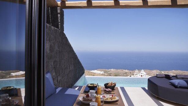 A balcony with a dining setup overlooking a scenic view of the sea and a white building. - Brilliance Suites - Adults Only (Santorini)