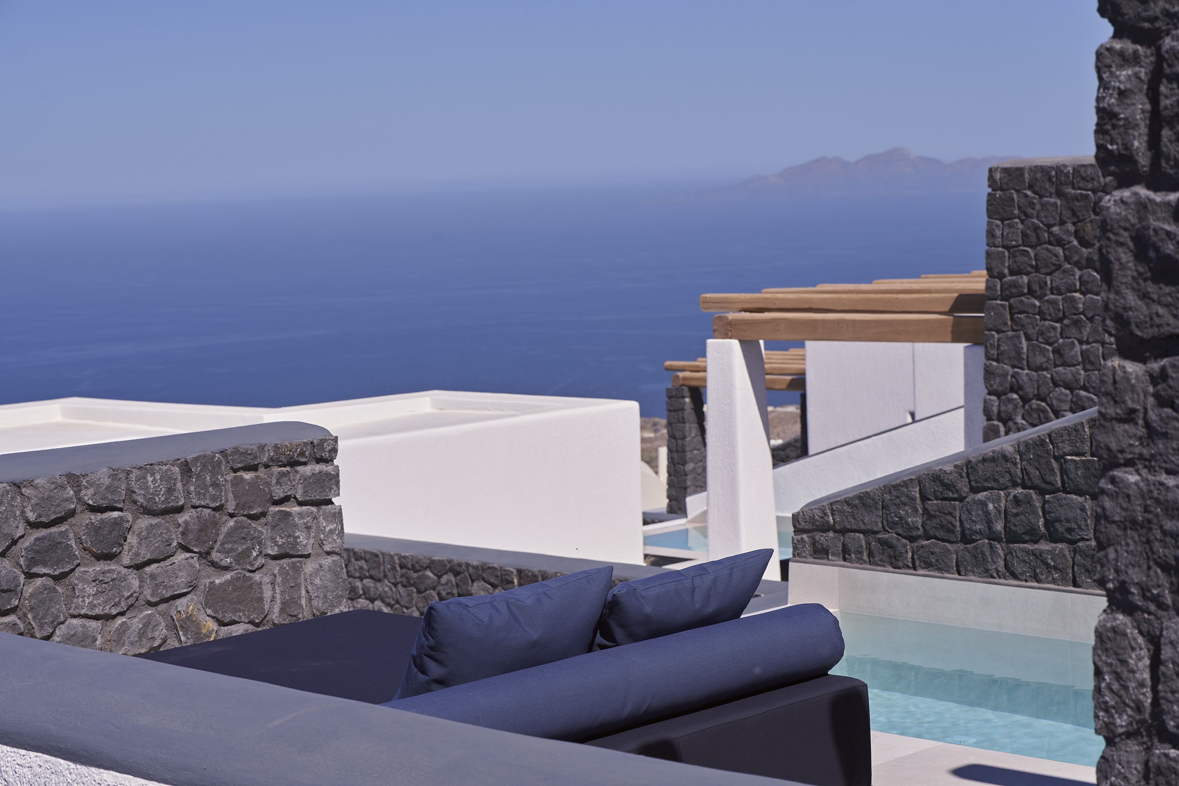 Un área de estar moderna al aire libre con pared de piedra y piscina, con vistas al mar azul turquesa y montañas lejanas.