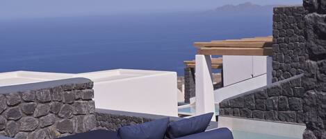Un área de estar moderna al aire libre con pared de piedra y piscina, con vistas al mar azul turquesa y montañas lejanas.