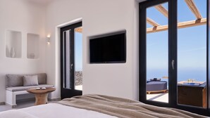 A bedroom with a large bed, a wooden bench, a small table, a TV, and a balcony with a view. - Brilliance Suites - Adults Only (Santorini)