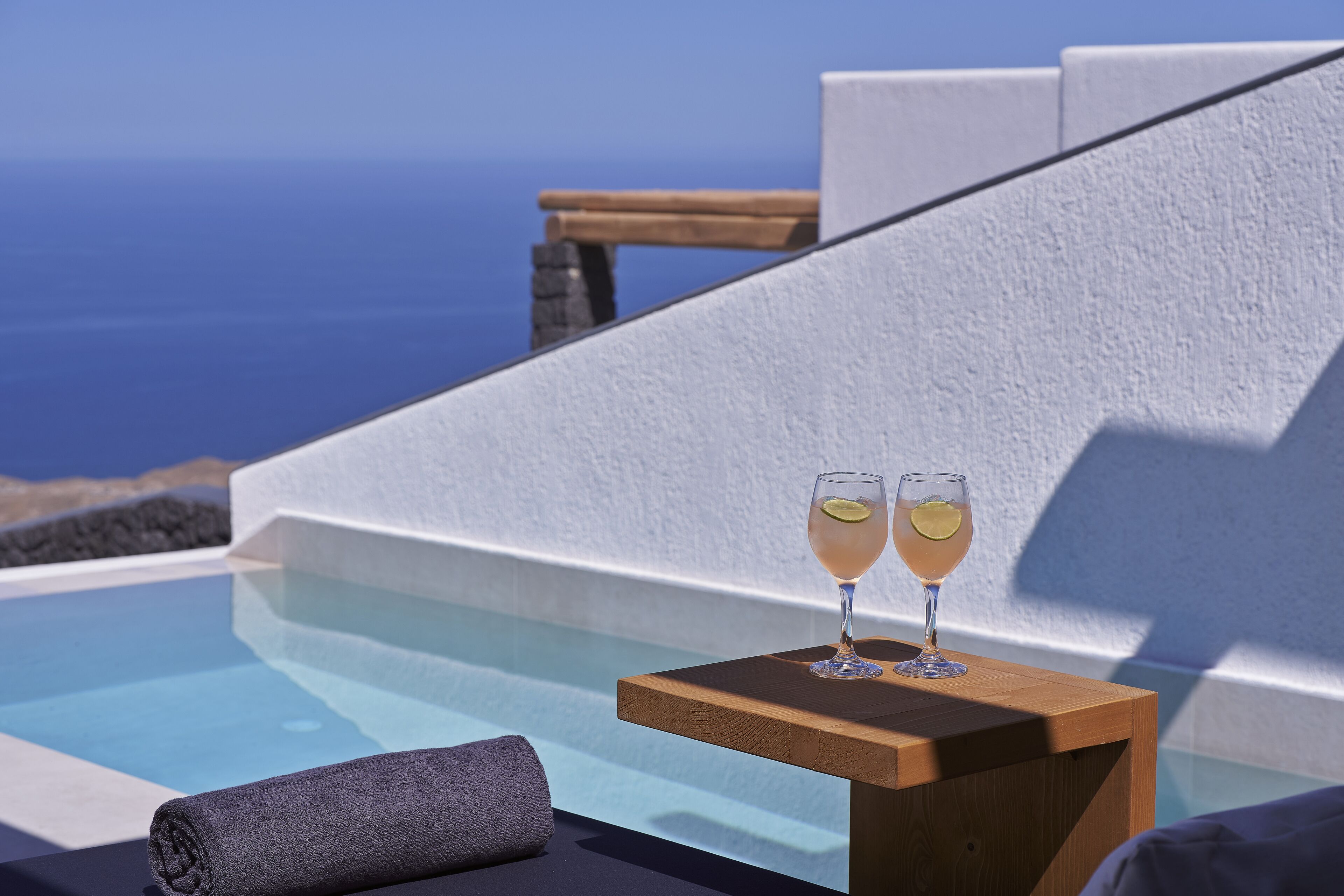 A poolside setting with two glasses of a light-colored beverage, a wooden table, and a view of the sea.
