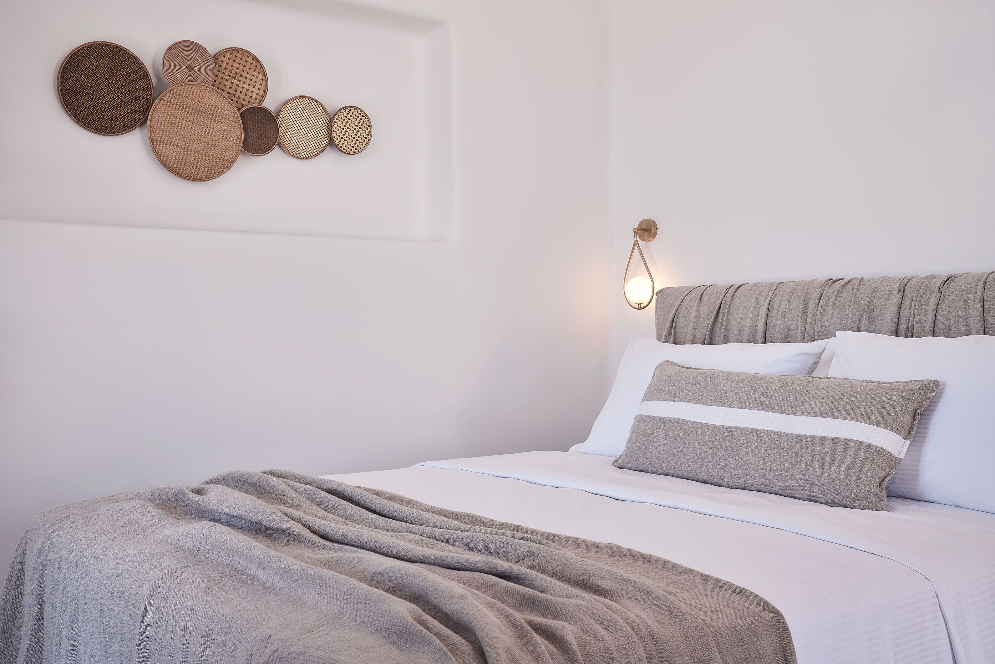 Una cama bien hecha con una manta gris, un cabecero con una lámpara y una pared decorada con un adorno circular.