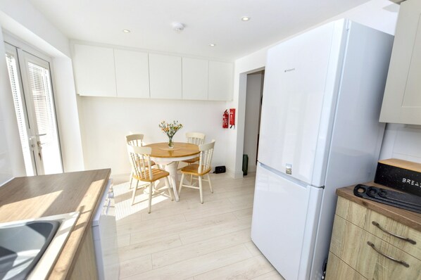 A modern kitchen with white cabinets, a wooden dining table with chairs, a refrigerator, and a small countertop with a sink. - A house that sleeps 4 guests in 2 bedrooms (Cromer)