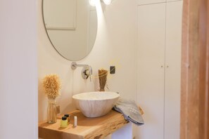 Une salle de bain avec un miroir rond, un meuble-lavabo en bois, un grand lavabo sur colonne et un porte-serviettes.